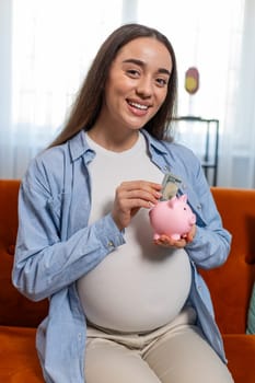 Happy thinking young pregnant woman inserting banknote in piggybank and caressing big belly sitting on sofa in living room at home. Smiling future mother lady on couch saving for baby's future concept