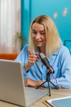 Happy blogger woman recording podcast at desk, giving workshop, webinar. Young Caucasian girl speaking at microphone and laptop webcam, making video call, discussing at home office in living room.
