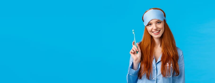 Health, beauty and lifestyle concept. Cheerful pretty european woman with blue eyes, toothy smile, holding toothbrush and smiling delighted, wearing sleep mask, nightwear, blue background.