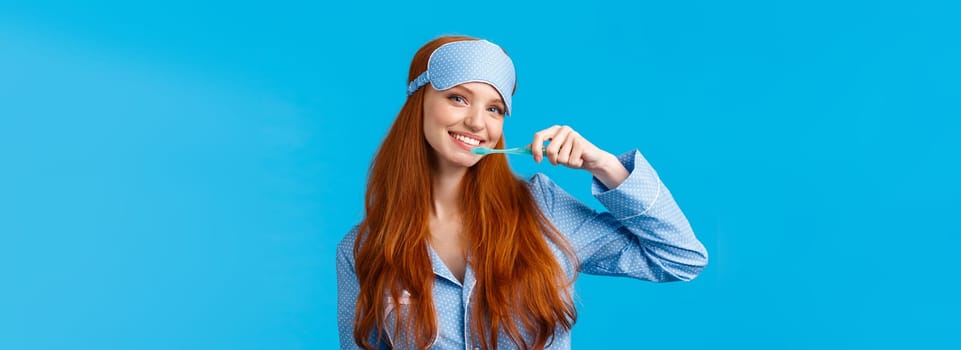 Cheerful cute redhead woman in nightwear, pyjama and sleep mask, brushing teeth holding toothbrush and smiling cheerful, wake up and taking care hygiene, standing blue background.