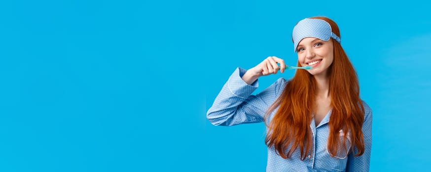 Cheerful cute redhead woman in nightwear, pyjama and sleep mask, brushing teeth holding toothbrush and smiling cheerful, wake up and taking care hygiene, standing blue background.