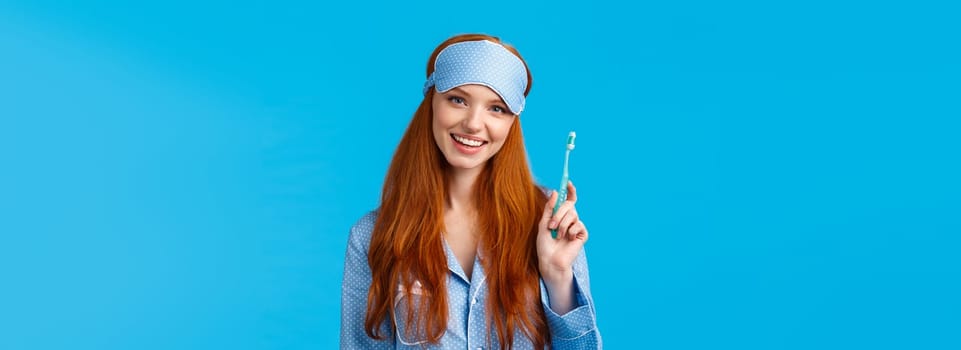 Health, beauty and lifestyle concept. Cheerful pretty european woman with blue eyes, toothy smile, holding toothbrush and smiling delighted, wearing sleep mask, nightwear, blue background.