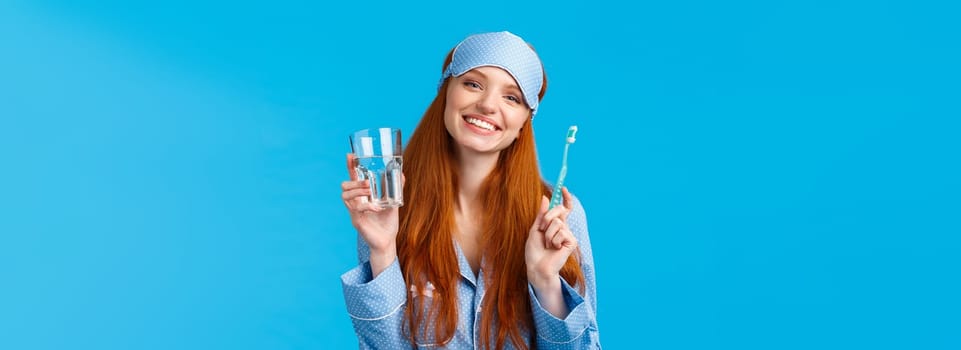 Happy smiling and carefree redhead enthusiastic girl in sleep mask, pyjama, holding glass water and toothbrush, taking care mouth hygiene, dental daily routine, blue background.
