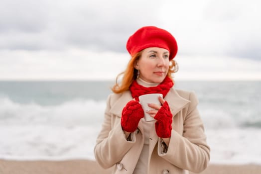 Woman beach in red beret scarf and mitts holding a cup of tea in his hands. Depicting beach relaxation and cozy attire. Walks by the sea.