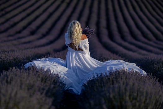 Blonde woman poses in lavender field at sunset. Happy woman in white dress holds lavender bouquet. Aromatherapy concept, lavender oil, photo session in lavender.