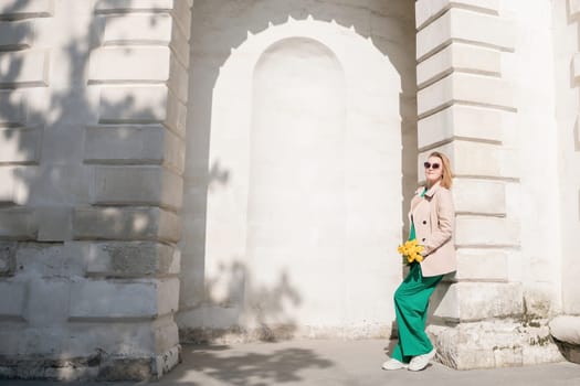 A woman is standing in front of a building with a yellow flower in her hand. She is wearing a green dress and a tan coat