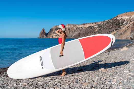 A woman in a red bikini and a Santa hat carries a SUP Board against the background of the sea, rocks. The concept of holiday and travel