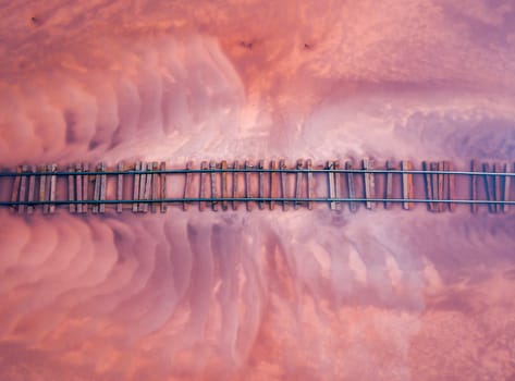 Aerial of a railroad tracks crossing a pink-hued lake. The perspective offers a unique view of the railway lines submerged in the shallow waters of the lake. Burlinskoe Lake, located in Bursol.