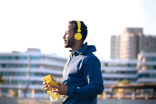 Athletic latin man with yellow headphones drinking water and looking at mobile phone on a break while exercising.