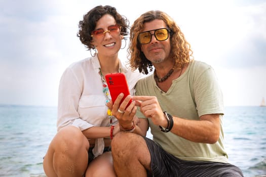 Middle aged man and woman sitting on the beach smiling browsing smartphone apps. Concept:Vacation and Technology
