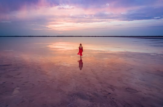 Aerial of a young woman in red dress walking in the water of a unique pink salt lake. Sunset at lake Bursol with beautiful reflections on calm water surface. Stunning scenery.