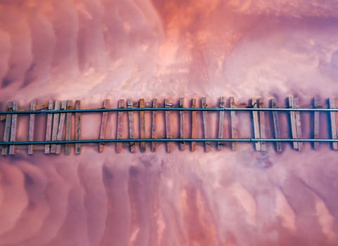 Aerial of a railroad tracks crossing a pink-hued lake. The perspective offers a unique view of the railway lines submerged in the shallow waters of the lake. Burlinskoe Lake, located in Bursol.