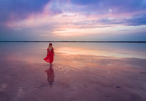 Aerial of a young woman in red dress walking in the water of a unique pink salt lake. Sunset at lake Bursol with beautiful reflections on calm water surface. Stunning scenery.