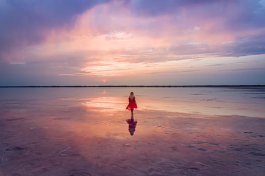 Aerial of a young woman in red dress walking in the water of a unique pink salt lake. Sunset at lake Bursol with beautiful reflections on calm water surface. Stunning scenery.