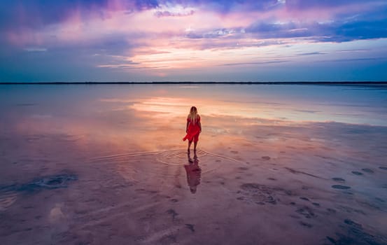 Aerial of a young woman in red dress walking in the water of a unique pink salt lake. Sunset at lake Bursol with beautiful reflections on calm water surface. Stunning scenery.