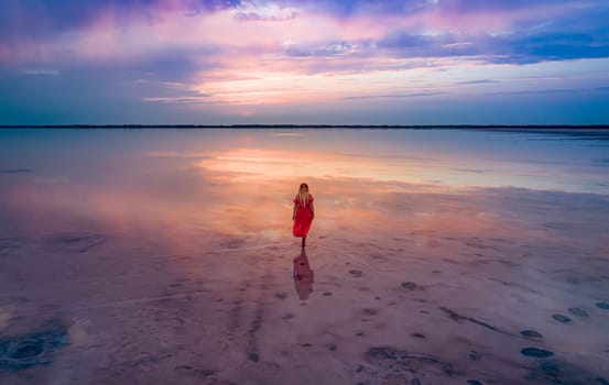 Aerial of a young woman in red dress walking in the water of a unique pink salt lake. Sunset at lake Bursol with beautiful reflections on calm water surface. Stunning scenery.