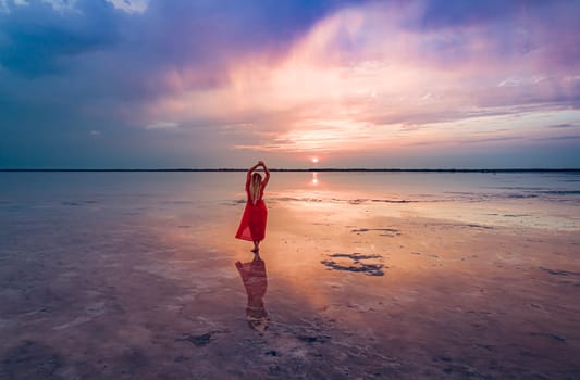 A woman in a red dress stands on the shallow waters of Burlinskoe Lake as the sun sets over the horizon, creating a beautiful reflection on the water.