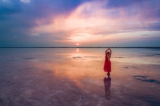 A woman in a red dress stands on the shallow waters of Burlinskoe Lake as the sun sets over the horizon, creating a beautiful reflection on the water.