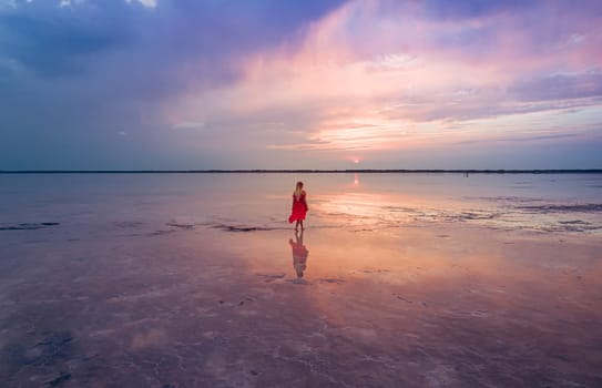 Aerial of a young woman in red dress walking in the water of a unique pink salt lake. Sunset at lake Bursol with beautiful reflections on calm water surface. Stunning scenery.