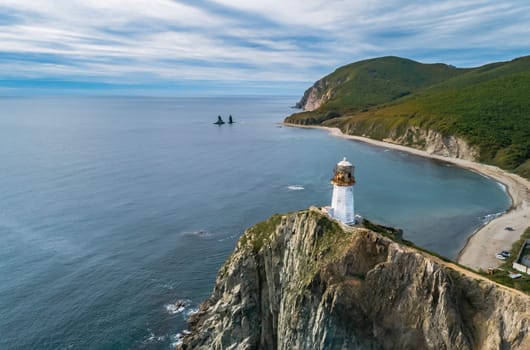 A panoramic aerial view captures the Rudnaya Pristan Lighthouse standing tall on a rugged cliff overlooking a picturesque bay.