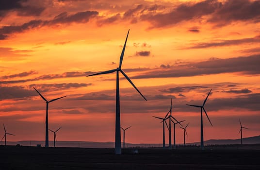 Multiple wind turbines silhouetted against a vibrant sunset sky. The turbines stand tall against the backdrop of orange and red clouds.