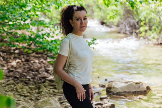 woman with a backpack on a hike stands by the river in a forest of green trees