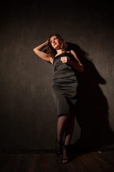 woman in a black dress stands in a dark room against the wall