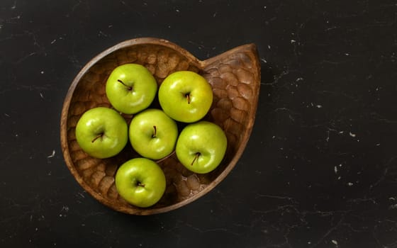 Top down view, six green apples in wooden carved bowl, on black marble like board.