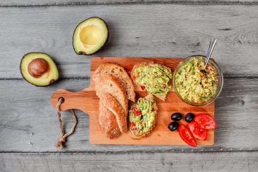 Flat lay photo, freshly prepared guacamole in small glass bowl, bread, tomatoes, olives at working board and two avocados next gray wood desk.