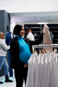 Smiling asian pregnant woman examining jacket on hanger while shopping for maternity clothes in mall. Expectant mother checking outfit on hanger while browsing rack with apparel