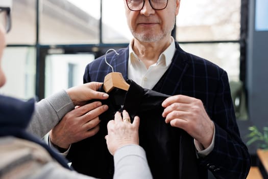 Elderly wife helping stylish husband choosing black shirt in modern boutique, looking to buy formal wear. Senior couple buying fashionable clothes and trendy merchandise in clothing store. Close up