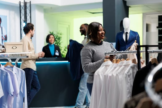Clothing store smiling customer holding jacket on hanger and checking size options in smartphone app. African american woman choosing formal outfit and using mobile phone website