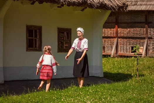 In front of a thatched building, a woman and a little girl are standing together. The scene includes elements like grass, a house, and people enjoying nature in the summertime