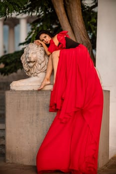 A woman in a red dress is sitting on a stone lion statue. The statue is white and has a lion's head. The woman is wearing a black belt and is looking at the camera. The scene has a calm