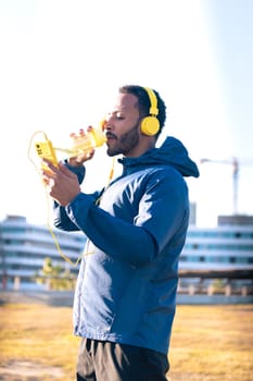 Athletic latin man with yellow headphones drinking water and looking at mobile phone on a break while exercising.