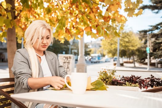 A blonde woman sits at a table with a cup of coffee and a leaf on it. The scene is set in a city with a tree in the background