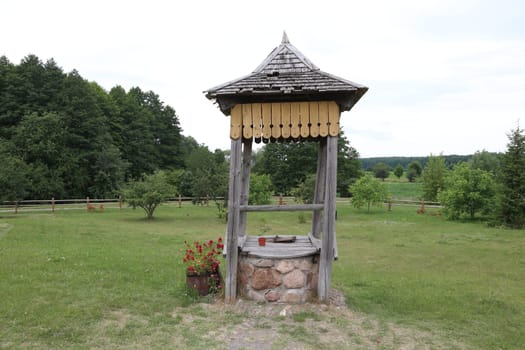 Decorative made of stone and wood, the base is made of small stones. The roof is wooden, against the background of green grass, forest, sky.