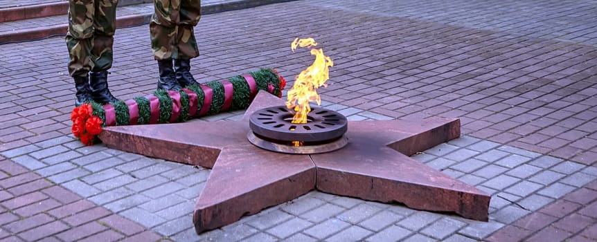 A soldier in camouflage uniform and boots stands at a monument with an eternal flame. A wreath and flowers honor the fallen. The soldier symbolizes dedication and respect.