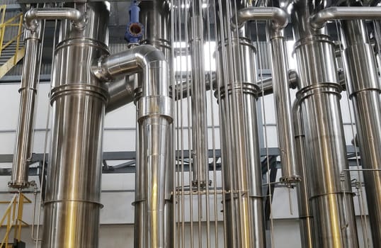 A chemical plant features a complex network of stainless steel pipes and glass vessels, creating an intricate system of interconnected straight and curved components in various sizes and shapes.