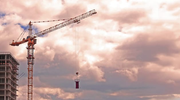 A construction crane stands tall against a backdrop of soft pink and white clouds, with a building under construction visible in the foreground.