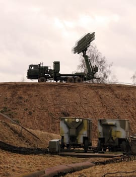 Two large military radar systems, mounted on vehicles, stand on a hill under a cloudy sky.