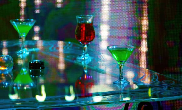 Three cocktails, two green and one red, sit on a glass table under a colorful neon light. The scene is vibrant and inviting.