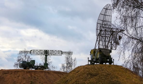Two large military radar systems, mounted on vehicles, stand on a hill under a cloudy sky.