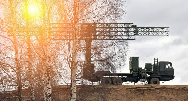 Truck-mounted military radar system silhouetted against setting sun with large complex dish and parked on dirt road with trees in background.