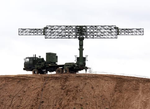 A large, military radar system is mounted on a truck and positioned on a dirt hilltop. The radar dish is extended and pointed upwards, likely scanning for potential threats in the sky.