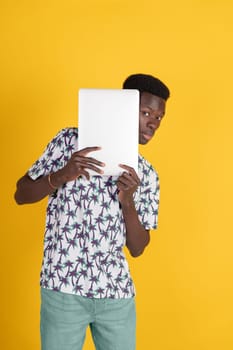 A man is holding a white laptop and looking at the camera. The image has a bright yellow background and a tropical print on the shirt. The man's expression is serious and focused