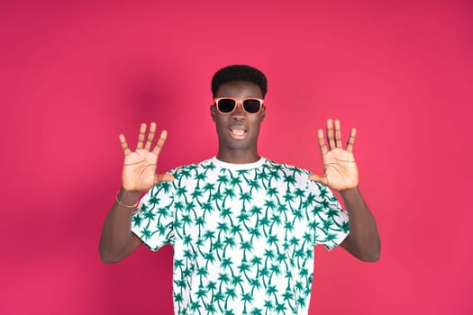 A man wearing star-patterned shirt and sunglasses stands against a pink backdrop, hands up signaling stop.