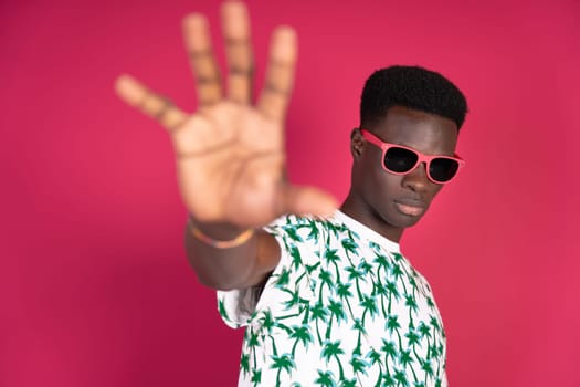 A man wearing sunglasses and a shirt with palm trees on it is standing in front of a pink background