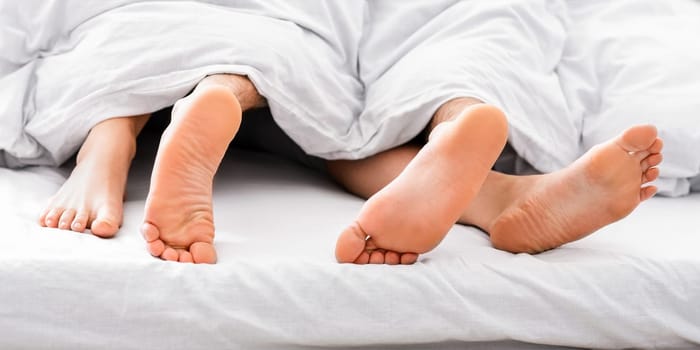 This image shows a close-up view of two bare feet sticking out from under white bedding. The feet appear to belong to two people, possibly a couple, lying in bed together