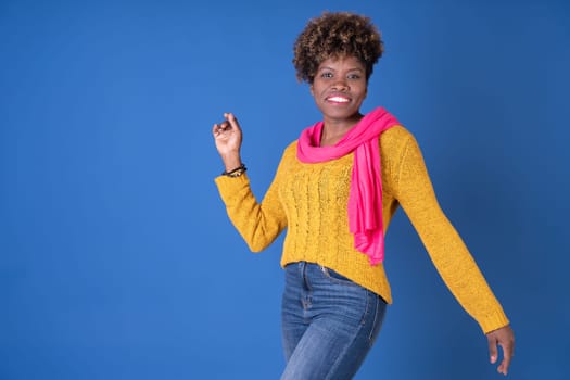 Young pretty black woman dancing with afro curly hair smiling in fuchsia scarf and yellow sweater on blue background with copy space. Happiness concept.